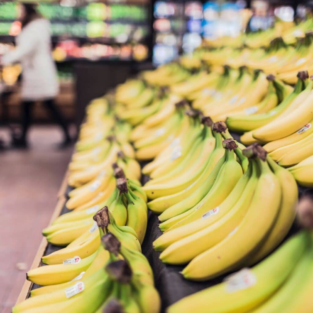Retail Grocery - An image of a rack of bananas with refrigerated goods stocked in the background and a lady pushing a shopping cart.