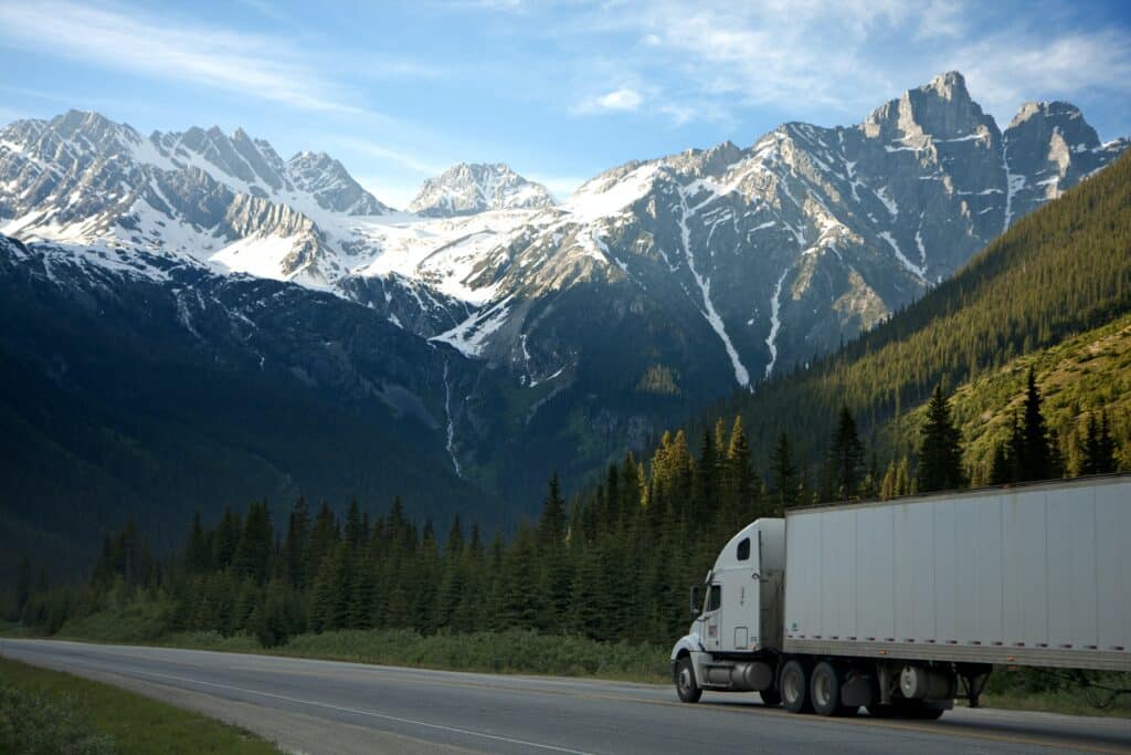 A tractor trailer represent a dry LTL truckload driving on a highway with snow-capped mountains in the background.